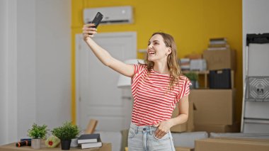 Genç sarışın kadın yeni evinde gülümseyen akıllı telefonuyla selfie çekiyor.