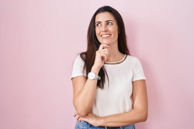 Young brunette woman standing over pink background with hand on chin thinking about question, pensive expression. smiling and thoughtful face. doubt concept. 