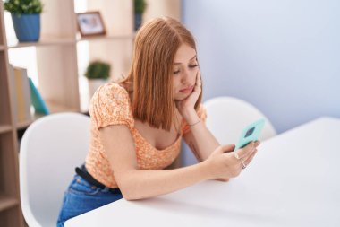 Young redhead woman using smartphone sitting on table at home