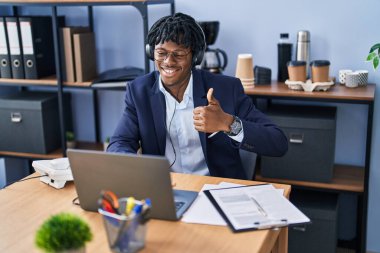 Ofiste çalışan Afrikalı genç adam kulaklık takıyor, gülümsüyor, gülümsüyor ve olumlu bakıyor. 