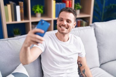 Genç ispanyol adam evdeki kanepede oturarak akıllı telefonuyla selfie çekiyor.