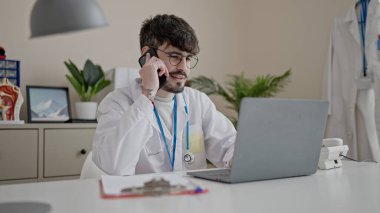 Genç İspanyol doktor bilgisayarını kullanarak klinikte telefonla konuşuyor.