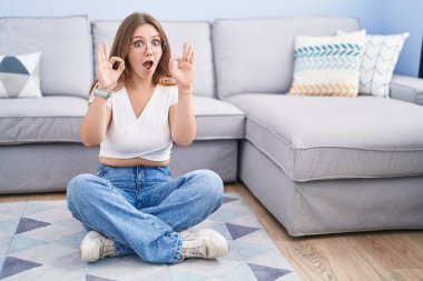 Beyaz bir kadın oturma odasında yerde oturuyor. Şaşırmış ve şok olmuş bir şekilde parmaklarıyla onay sembolü yapıyor. çılgın ifade 