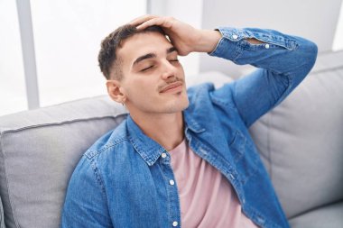 Young hispanic man relaxed with hand on head sitting on sofa at home