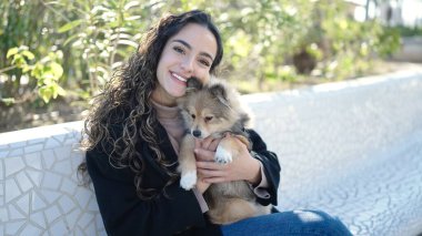 Köpeğiyle gülümseyen genç İspanyol kadın parkta bankta oturuyor.