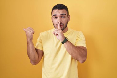 Young hispanic man standing over yellow background asking to be quiet with finger on lips pointing with hand to the side. silence and secret concept. 