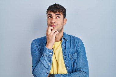 Young hispanic man standing over blue background with hand on chin thinking about question, pensive expression. smiling with thoughtful face. doubt concept. 