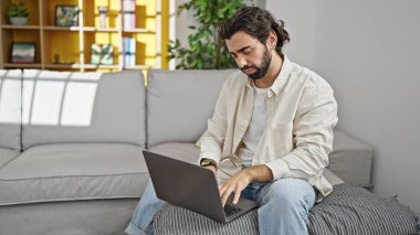 Genç İspanyol adam evdeki koltukta dizüstü bilgisayar kullanıyor.