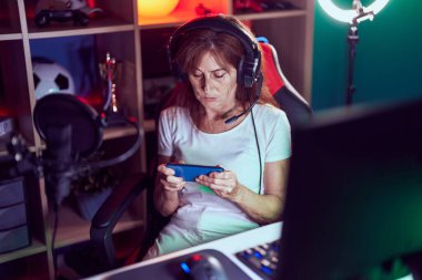 Middle age woman streamer playing video game using smartphone at gaming room