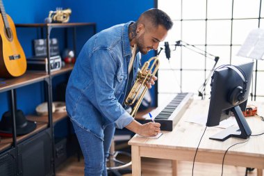 Young hispanic man musician composing song holding trumpet at music studio