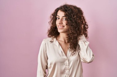 Hispanic woman with curly hair standing over pink background smiling with hand over ear listening an hearing to rumor or gossip. deafness concept. 