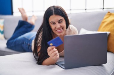 Evdeki kanepede dizüstü bilgisayar ve kredi kartı kullanan genç ve güzel bir İspanyol kadın.