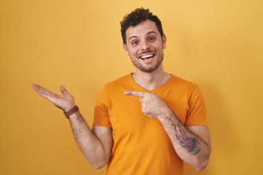 Young hispanic man standing over yellow background amazed and smiling to the camera while presenting with hand and pointing with finger. 