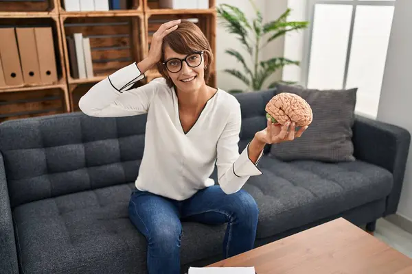 Brunette woman working at therapy office holding brain stressed and frustrated with hand on head, surprised and angry face