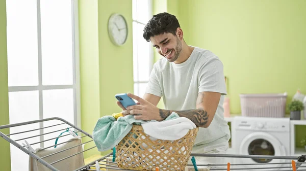 在洗衣房用智能手机靠在篮子上穿衣服的年轻人 — 图库照片