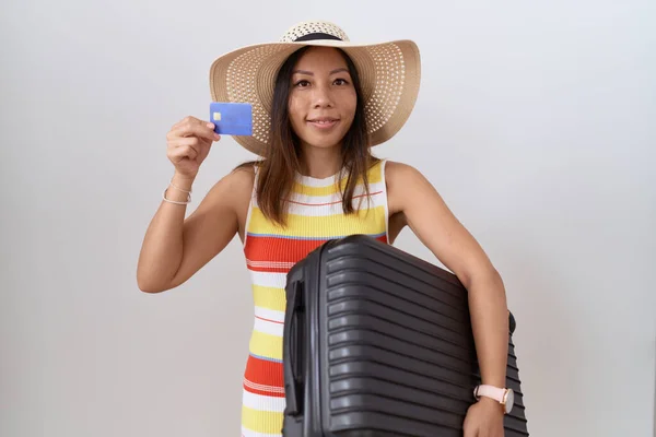 Middle age chinese woman holding suitcase and credit card smiling with a happy and cool smile on face. showing teeth.