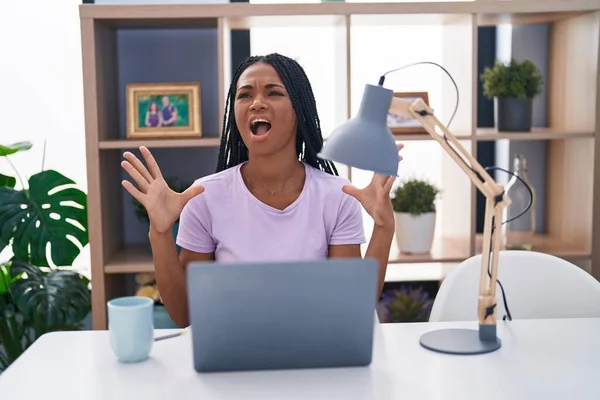 stock image African american woman with braids using laptop at home crazy and mad shouting and yelling with aggressive expression and arms raised. frustration concept. 