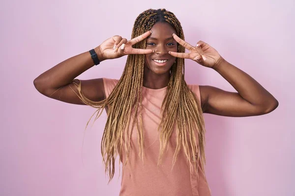 非洲裔美国妇女 辫子式的 站在粉红的背景上 用手指捂住脸 愉快地微笑着 表示胜利 — 图库照片