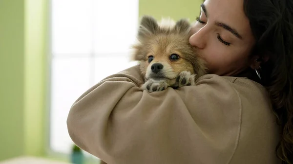 Young hispanic woman with dog hugging and kissing standing at dinning room