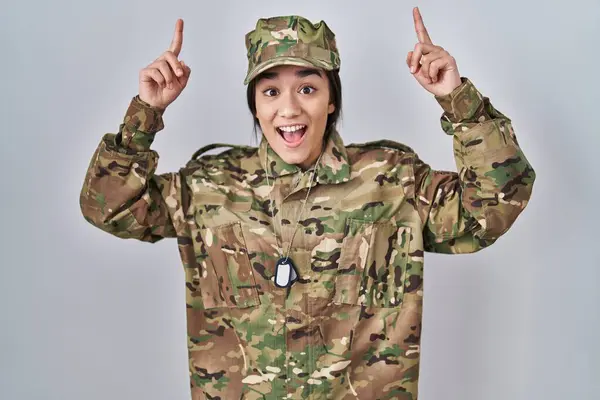 Young south asian woman wearing camouflage army uniform smiling amazed and surprised and pointing up with fingers and raised arms.
