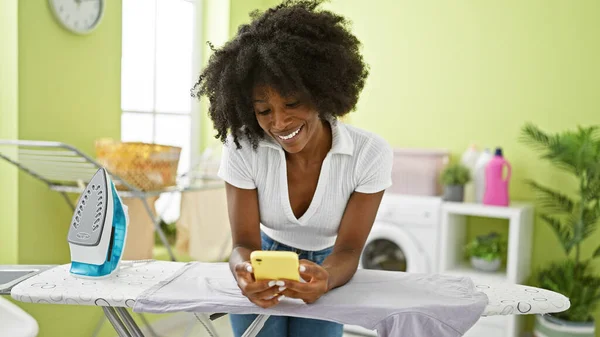 African american woman talking on smartphone leaning on ironing machine at laundry room