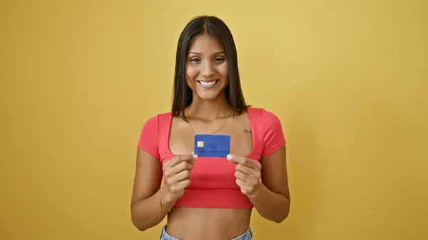 年轻的拉丁女人带着自信的笑容 在孤立的黄色背景下拿着信用卡 — 图库照片