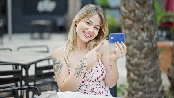 Young blonde woman tourist pointing to credit card smiling at coffee shop terrace