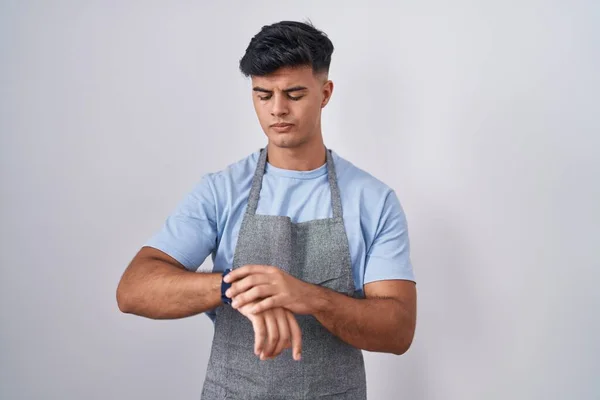 Hispanic young man wearing apron over white background checking the time on wrist watch, relaxed and confident
