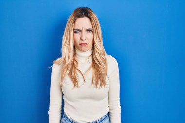 Mavi arka planda duran genç beyaz kadın şüpheci ve gergin, sorun yüzünden surat asıyor. negatif kişi. 