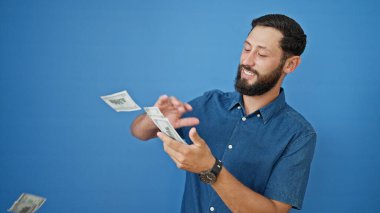 Genç İspanyol adam gülümsüyor kendinden emin, izole edilmiş mavi arka plana dolar atıyor.