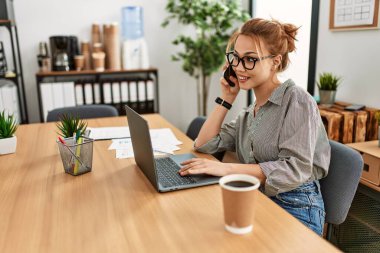 Kafkasyalı genç bir iş adamı bilgisayarını kullanarak ofisteki akıllı telefondan konuşuyor.