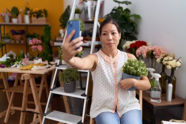 Genç Asyalı kadın çiçekçi çiçekçide akıllı telefon fabrikasıyla selfie çekiyor.