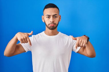 Mavi arka plan üzerinde duran genç İspanyol adam üzgün ve üzgün görünüyor, parmaklarıyla yön gösteriyor, mutsuz ve depresif.. 