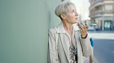 Orta yaşlı sarışın kadın gülümsüyor. Kendine güveniyor. Akıllı telefonuyla sokakta sesli mesaj yolluyor.