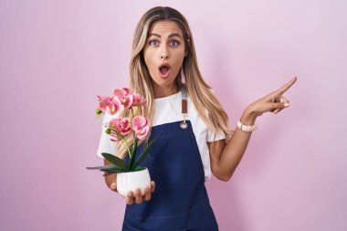 Bahçıvan önlüğü giyen genç sarışın kadın elinde bitki tutarken şaşkın bir şekilde parmağıyla yüzünü işaret etti, ağzı açık hayret içinde kaldı.. 