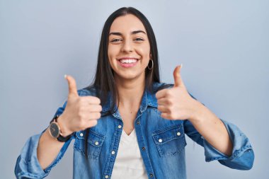 Mavi arka plan başarı işaretinin üzerinde duran İspanyol kadın elleriyle olumlu jest yapıyor, gülümsüyor ve mutlu. Neşeli ifade ve kazanan jest. 
