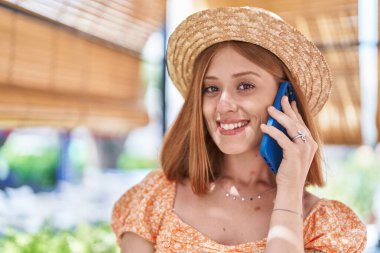 Young redhead woman tourist wearing summer hat talking on smartphone at street