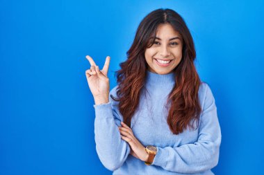 Mavi arka planda dikilmiş, gülümseyen, kameraya göz kırpan, parmaklarıyla zafer işareti yapan İspanyol genç bir kadın. İki numara.. 