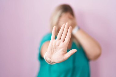 Beyaz tenli kadın pembe arka planda duruyor ellerle gözleri örtüyor ve üzgün ve korku ifadesiyle dur hareketi yapıyor. utangaç ve olumsuz kavram. 