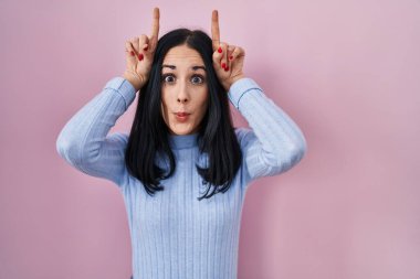 İspanyol kadın pembe arka planda dikilmiş, parmaklarıyla boynuz gibi komik hareketler yapıyor. 