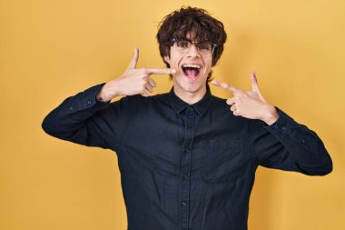 Sarı arka planda gözlük takan genç adam gülümseyip, parmaklarıyla ve ağzıyla işaret ediyor. diş sağlığı kavramı. 