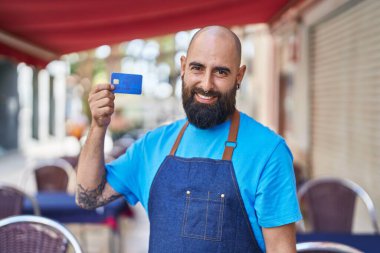 Young bald man waiter smiling confident holding credit card at coffee shop terrace