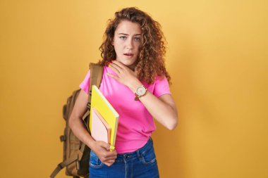 Young caucasian woman wearing student backpack and holding books cutting throat with hand as knife, threaten aggression with furious violence 