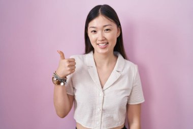 Chinese young woman standing over pink background doing happy thumbs up gesture with hand. approving expression looking at the camera showing success. 
