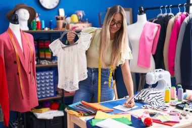 Young beautiful hispanic woman tailor holding t shirt writing on notebook at clothing factory