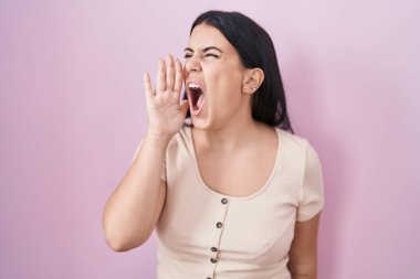 Young hispanic woman standing over pink background shouting and screaming loud to side with hand on mouth. communication concept. 
