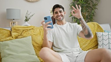 Young hispanic man using smartphone doing ok gesture at home