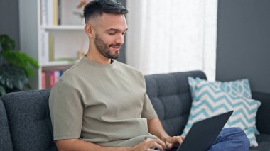 Genç İspanyol adam evdeki koltukta dizüstü bilgisayar kullanıyor.