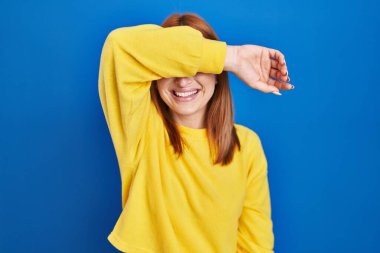 Mavi arka planda duran genç bir kadın. Gözleri kollarıyla kaplıyor. Neşeli ve komik gülümsüyor. kör kavram. 