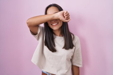 Pembe arka planda duran genç İspanyol kadın kollarıyla gözlerini kapatıyor neşeli ve komik gülümsüyor. kör kavram. 